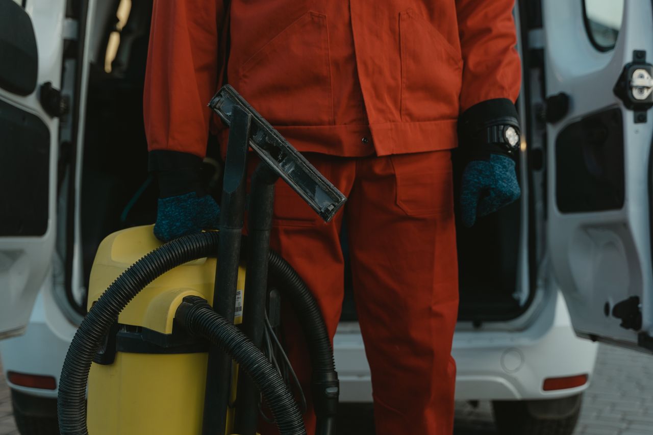 Une technicienne de nettoyage professionnelle en uniforme rouge tenant un aspirateur près d’un van, prête à intervenir.